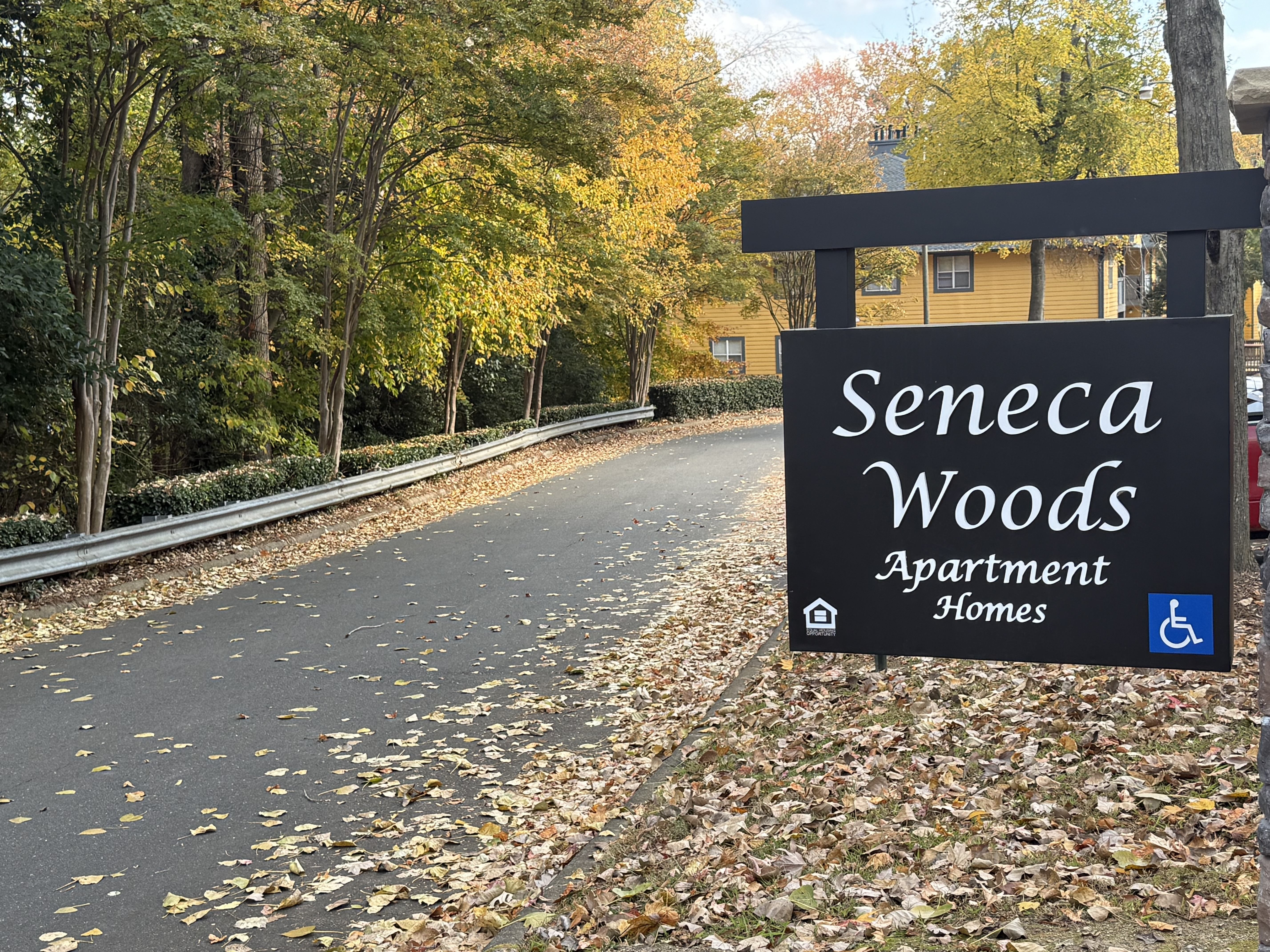 A sign for Seneca Woods Apartment Homes is shown in front of a tree-lined street.